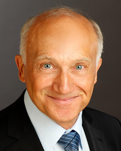 Man in dark suit and white shirt with blue tie against a dark background looks at the camera with a friendly smile.