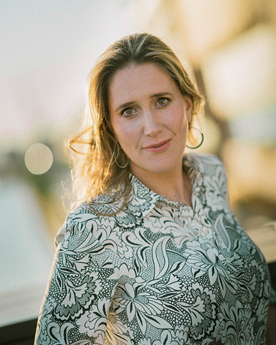 Woman in floral-patterned shirt looks into the camera with a friendly expression, her hair glowing in the light of the sun behind her